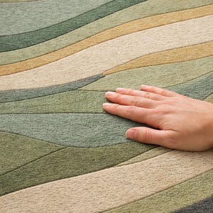 Pu&ograve; includere: Primo piano di un tappeto con un motivo a onde nelle tonalit&agrave; del verde, beige e marrone. Il tappeto ha un aspetto morbido e strutturato. Una mano &egrave; appoggiata sul tappeto, evidenziando la sua consistenza e le variazioni di colore.
