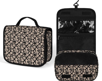 Op de afbeelding: Een zwarte toilettas met beige bloemenprint, een ritssluiting en een ophanghaak. De tas heeft meerdere vakken en een transparant plastic venster.