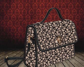 Può includere: Una borsa a mano nera di tipo cartella con un motivo floreale bianco e una chiusura dorata. La borsa ha una tracolla lunga regolabile e un manico superiore.