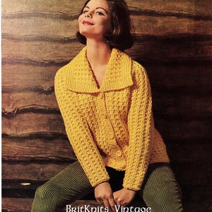 May include: A yellow knitted cardigan with a wide collar and button closure. The cardigan is worn by a model with short brown hair and is photographed against a wood panel background.