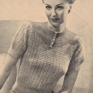 May include: A vintage-style, short-sleeved sweater with a textured, grid-like pattern. The sweater features a high, ribbed collar with three buttons and is likely made of a soft, light-colored yarn. The image is a black and white photograph.