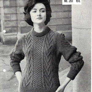 May include: A black and white photograph of a woman wearing a grey knitted jumper with a cable knit pattern. The jumper has a round neckline and long sleeves. The photo is from a vintage knitting pattern book and includes the text "No. 261 6d."