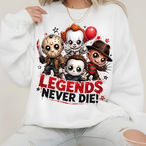 May include: A white long-sleeve sweatshirt featuring a graphic print of four chibi-style horror movie characters. Below the characters, the text reads "LEGENDS NEVER DIE!" in bold red and black lettering, accented by small black and red stars.