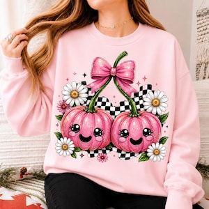 May include: A light pink crewneck sweatshirt featuring a graphic print of two smiling, glittery pink pumpkins with green stems tied together by a large pink bow. The design includes white daisies, green leaves, and a black and white checkered pattern.
