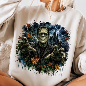 May include: A cream-coloured crewneck sweatshirt featuring a graphic print of Frankenstein's monster with outstretched hands. The design includes a dark castle, lightning, and scattered orange autumn leaves against a blue and black watercolour splash.