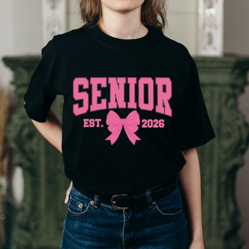 May include: Black t-shirt with the word "SENIOR" in pink block letters. Below the word "SENIOR" is a pink bow graphic, with the text "EST. 2026" in pink. The shirt is worn with blue jeans.