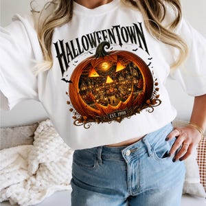 May include: A white t-shirt featuring a large graphic of a carved jack-o'-lantern pumpkin. Inside the pumpkin is a detailed scene of a town at night. The text HALLOWEENTOWN is printed above the pumpkin, and EST. 1998 is printed below it.