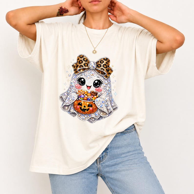 May include: An off-white short-sleeve t-shirt featuring a graphic of a cartoon ghost with large black eyes, a leopard print bow, and a smiling expression. The ghost is holding a small orange jack-o'-lantern filled with colourful candy pieces.