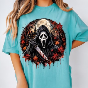 May include: A turquoise t-shirt featuring a circular graphic of a hooded figure with a white ghost mask holding a knife. The background shows a dark house, a full moon, and red autumn leaves. The bottom of the circular design has a dripping effect.