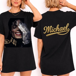 May include: Black t-shirt featuring a glittery image of Michael Jackson on the front. The image shows Jackson wearing a glove and smiling. The back of the shirt has the name "Michael" in gold script. A classic tee for music fans.
