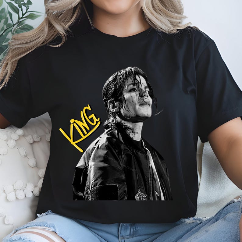 May include: Black t-shirt featuring a black and white image of a person looking upwards. The word "KING" is written in yellow, stylized text. The shirt is a crew neck style.