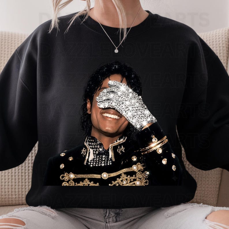 May include: Black sweatshirt featuring a smiling image of Michael Jackson wearing a sparkling glove and a black jacket with gold accents. The image is a close-up of the singer, with his hand covering his eyes.