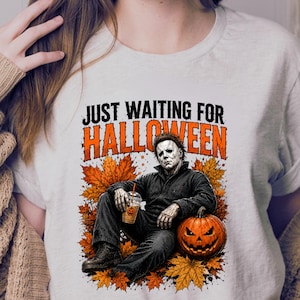 May include: A light-coloured t-shirt featuring a graphic of a character in a dark jumpsuit sitting among orange autumn leaves. The character holds a plastic cup with a pumpkin icon. The text reads "JUST WAITING FOR HALLOWEEN" in black and orange.