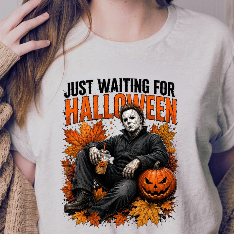 May include: A light-coloured t-shirt featuring a graphic of a character in a dark jumpsuit sitting among orange autumn leaves. The character holds a plastic cup with a pumpkin icon. The text reads "JUST WAITING FOR HALLOWEEN" in black and orange.