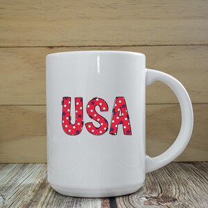 Puede incluir: Taza de cer&aacute;mica blanca con un asa grande. La taza presenta la palabra "USA" en letras rojas rellenas de estrellas blancas sobre un fondo azul. La taza est&aacute; sobre una superficie de madera.