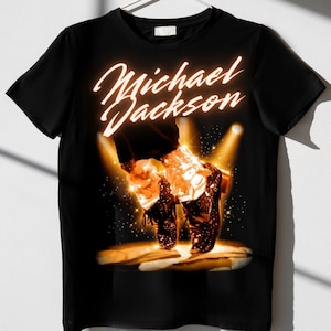 May include: Black t-shirt featuring a graphic of Michael Jackson's feet performing the moonwalk, illuminated by stage lights. The name "Michael Jackson" is written in a glowing, cursive font above the image. A classic design for music fans.