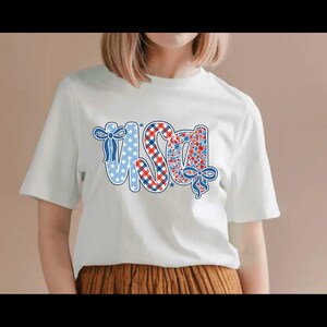Puede incluir: Camiseta blanca con la palabra "USA" en un dise&ntilde;o patri&oacute;tico. Las letras est&aacute;n decoradas con estrellas azules, cuadros rojos y blancos y un estampado floral. Lazos azules acent&uacute;an las letras.