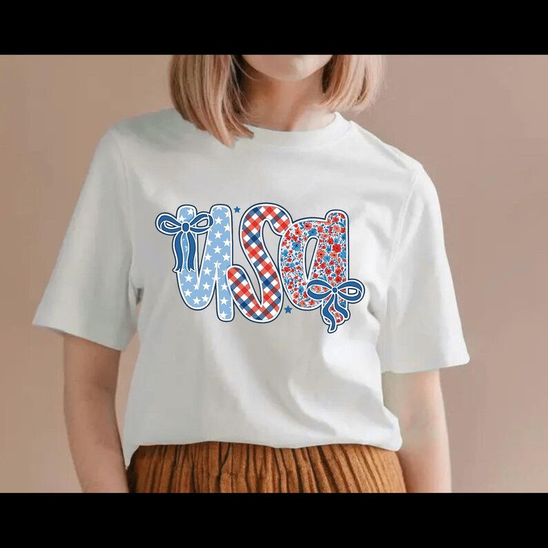 Puede incluir: Camiseta blanca con la palabra "USA" en un dise&ntilde;o patri&oacute;tico. Las letras est&aacute;n decoradas con estrellas azules, cuadros rojos y blancos y un estampado floral. Lazos azules acent&uacute;an las letras.