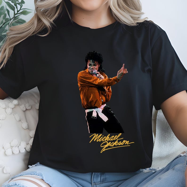May include: Black t-shirt featuring a vibrant image of Michael Jackson performing on stage. The image shows Jackson in a brown jacket and black pants, holding a microphone. The words "Michael Jackson" are written in gold script.