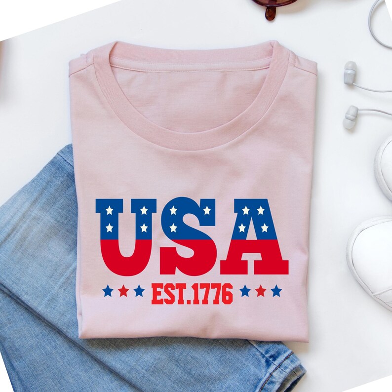 Puede incluir: Una camiseta rosa claro con la palabra "USA" en azul y rojo, con estrellas blancas. El texto "EST.1776" est&aacute; debajo de la palabra "USA". La camiseta est&aacute; doblada sobre unos vaqueros azules.