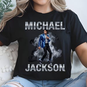 May include: Black t-shirt featuring a graphic of Michael Jackson in a blue sequined jacket and black pants, with the name "MICHAEL JACKSON" in silver block letters. The image includes a smoky background.