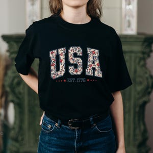Puede incluir: Camiseta negra con la palabra "USA" en un dise&ntilde;o floral con flores rojas y blancas. Debajo de la palabra "USA" est&aacute; el texto "EST. 1776" con estrellas a cada lado. La camiseta se combina con vaqueros azules.