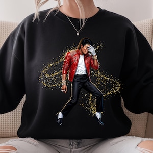 May include: Black sweatshirt featuring a graphic of a person in a red leather jacket, black pants, and white shirt. The person is wearing a white glove and striking a pose. Gold glitter-like accents surround the figure.