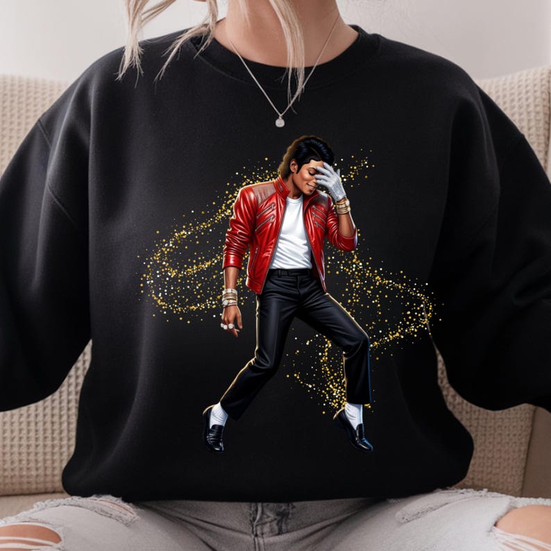 May include: Black sweatshirt featuring a graphic of a person in a red leather jacket, black pants, and white shirt. The person is wearing a white glove and striking a pose. Gold glitter-like accents surround the figure.