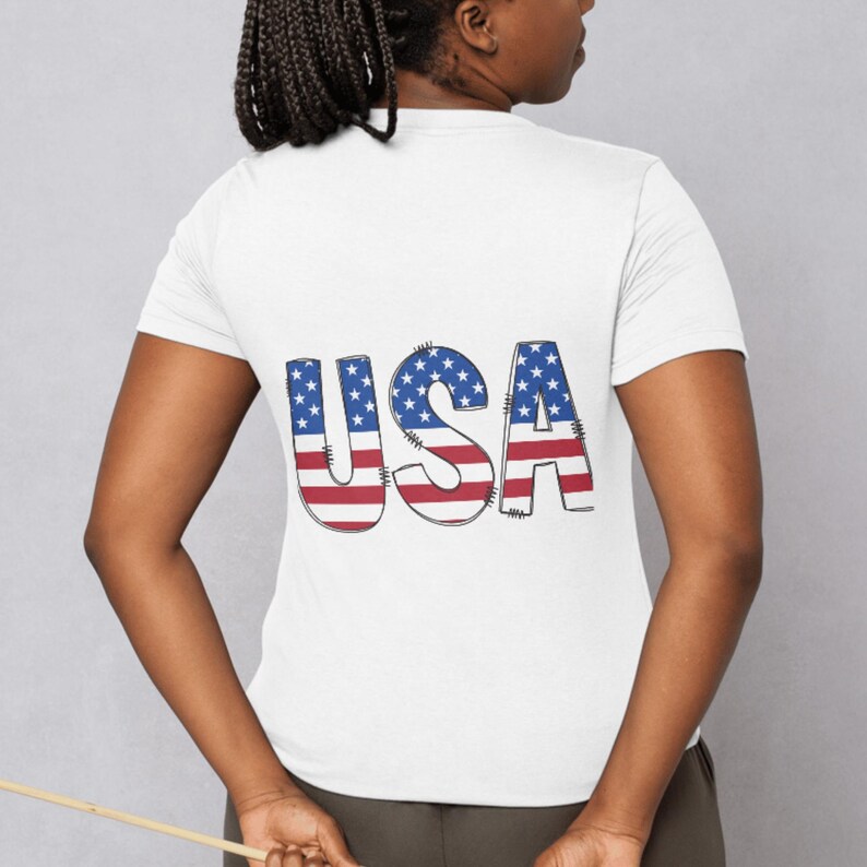 Puede incluir: Camiseta blanca con la palabra "USA" impresa en la espalda. Las letras est&aacute;n dise&ntilde;adas con un tema patri&oacute;tico, con estrellas azules y rayas rojas, que representan la bandera estadounidense. La camiseta tiene mangas cortas.