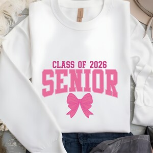May include: White sweatshirt with the text "CLASS OF 2026 SENIOR" in pink block letters and a pink bow graphic. The sweatshirt is a crew neck style.