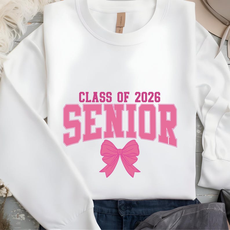 May include: White sweatshirt with the text "CLASS OF 2026 SENIOR" in pink block letters and a pink bow graphic. The sweatshirt is a crew neck style.