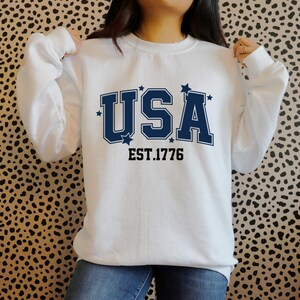 Puede incluir: Sudadera blanca de cuello redondo con la palabra "USA" en letras may&uacute;sculas azul marino, acentuada con estrellas. Debajo, el texto "EST. 1776" en negro. Un estilo casual y c&oacute;modo.