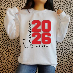 May include: White sweatshirt with the word "Senior" in black script and the numbers "2026" in red block letters. Five black stars are below the numbers. The sweatshirt is a crew neck style.