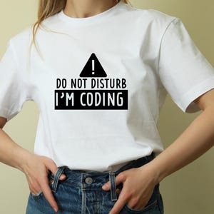May include: White t-shirt with black text that reads "Do Not Disturb I'm Coding" and a black warning sign with an exclamation point.