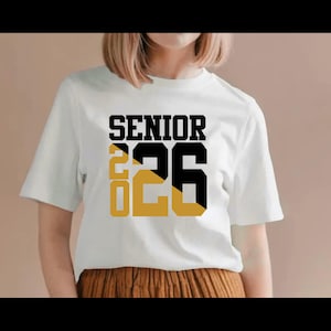 May include: White t-shirt with the word "SENIOR" in black block letters above the numbers "26" in black and gold. The number "20" is in gold. The shirt is short-sleeved.
