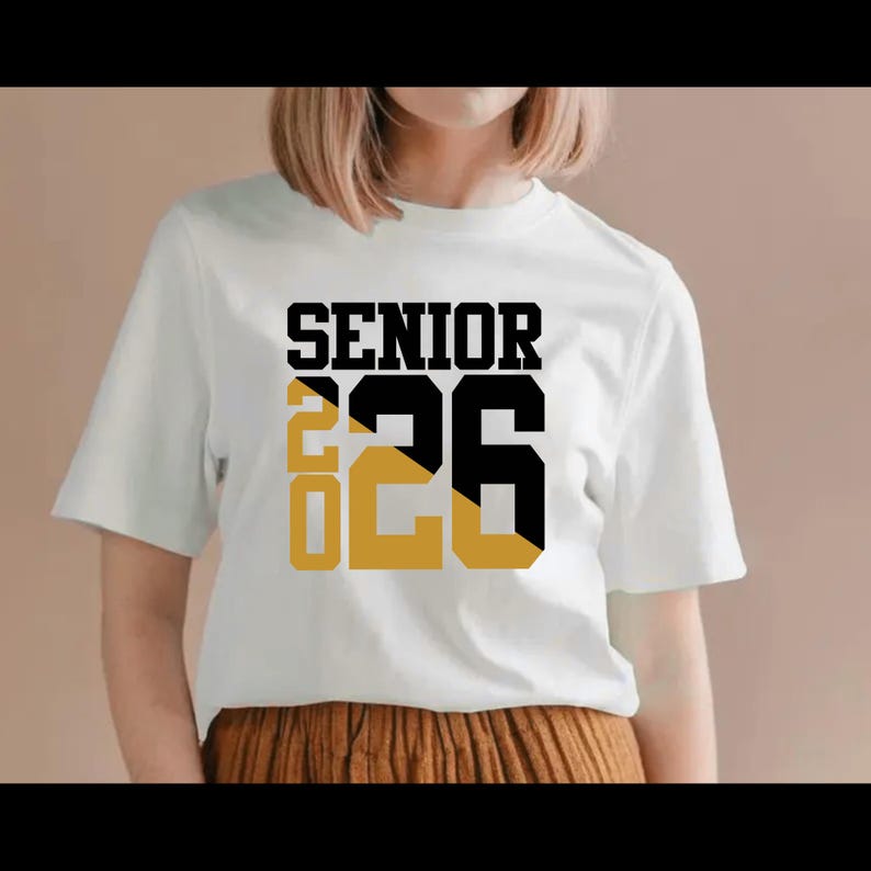 May include: White t-shirt with the word "SENIOR" in black block letters above the numbers "26" in black and gold. The number "20" is in gold. The shirt is short-sleeved.