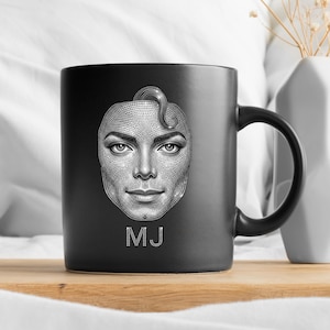 May include: Black ceramic mug featuring a detailed grayscale portrait of a person's face, with the initials "MJ" in white at the bottom. The mug has a black handle and is set against a neutral background.