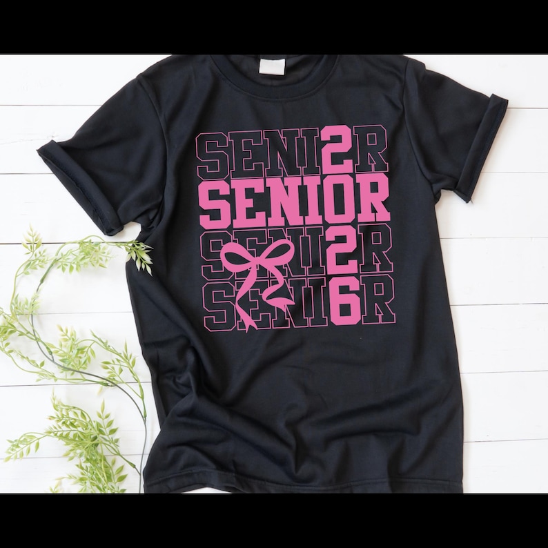 May include: Black t-shirt with the word "SENIOR" in pink, repeated four times, with a pink bow graphic. The shirt is made of a soft material and has short sleeves. The design is centered on the front of the shirt.