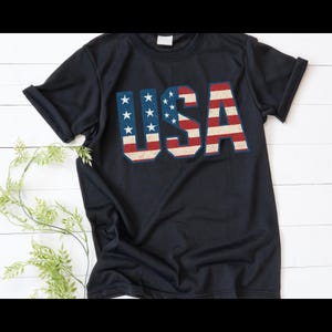 Puede incluir: Una camiseta azul marino con la palabra "USA" impresa en la parte delantera. Las letras est&aacute;n dise&ntilde;adas con un patr&oacute;n de bandera estadounidense desgastada, con rayas rojas y blancas y campos azules con estrellas blancas. La camiseta tiene mangas cortas.