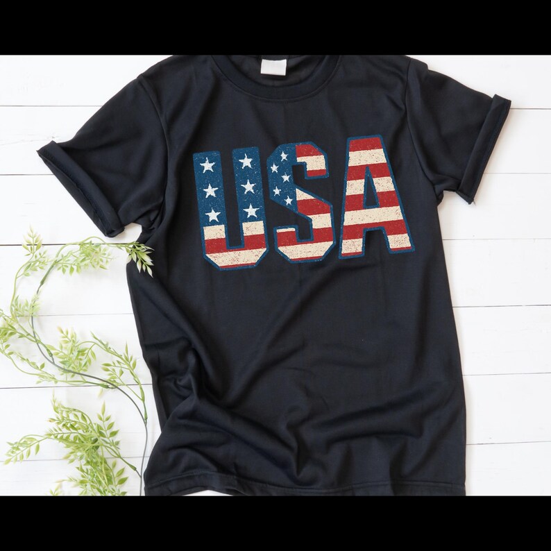 Puede incluir: Una camiseta azul marino con la palabra "USA" impresa en la parte delantera. Las letras est&aacute;n dise&ntilde;adas con un patr&oacute;n de bandera estadounidense desgastada, con rayas rojas y blancas y campos azules con estrellas blancas. La camiseta tiene mangas cortas.