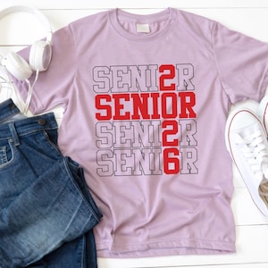 May include: A light purple t-shirt with the word "SENIOR" in red and black block letters. The word is repeated four times down the front of the shirt. A pair of blue jeans, white headphones, and white sneakers are also in the image.