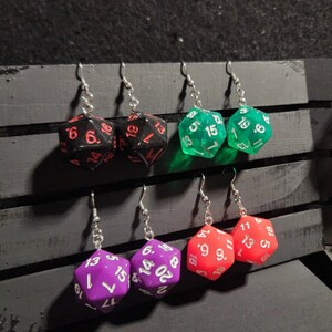 Peut inclure: Six paires de boucles d'oreilles pendantes avec une variété de couleurs et de motifs de dés. Les dés sont noirs, verts, violets et rouges. Les boucles d'oreilles sont en métal et ont des crochets argentés.