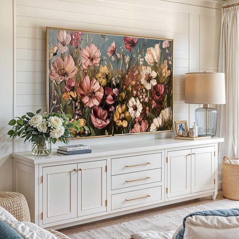 May include: A white dresser with three drawers and two double doors. The dresser has gold hardware. A large painting of pink and yellow flowers hangs above the dresser. A lamp with a beige shade and a clear glass base sits on the dresser.