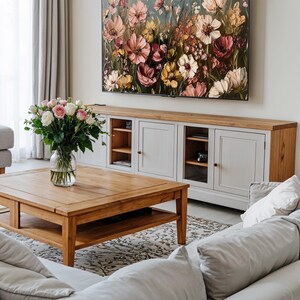 May include: A living room with a white sofa, a wooden coffee table, and a large floral painting hanging above a white cabinet. The painting features a variety of pink, yellow, and white flowers.