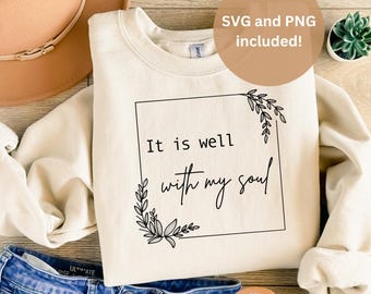 Puede incluir: Una sudadera de color crema con el texto "It is well with my soul" dentro de un marco cuadrado negro con detalles florales. La sudadera está colocada sobre una superficie de madera con un sombrero marrón, unos vaqueros azules y una planta en maceta.