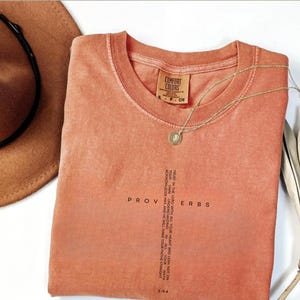 Peut inclure: Un t-shirt Comfort Colors de couleur corail avec le mot "PROVERBES" imprimé verticalement et le texte "TRUST IN THE LORD..." en forme de croix. Un collier en or avec un pendentif repose sur le t-shirt. Un chapeau marron est visible en arrière-plan.