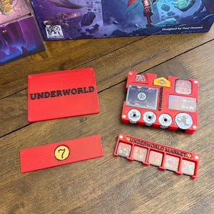 May include: Red game components with the word "UNDERWORLD" printed on them. The components include a rectangular piece with the number 7, and a red game board with the words "UNDERWORLD MARKET". A game box is visible in the background.