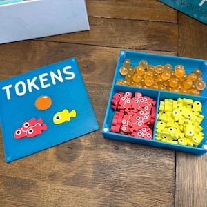 Puede incluir: Un organizador de fichas de juego azul con la palabra "TOKENS" en blanco. El organizador contiene piezas de juego naranjas, rojas y amarillas. Las fichas tienen forma de pez y son redondas. Las piezas del juego están en una bandeja azul.