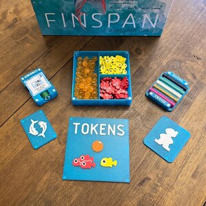 Könnte beinhalten: Ein Brettspiel namens "Finspan" mit verschiedenen Spielteilen. Dazu gehören ein blauer Behälter mit orangefarbenen, gelben und roten Spielsteinen, blaue Karten und eine blaue Box mit farbigen Stiften. Die Spielbox ist im Hintergrund zu sehen.