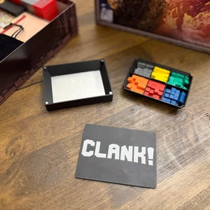 May include: A board game setup with various components on a wooden surface. The game box is partially visible, along with a black tray, a container with colored game pieces, and a card that reads "CLANK!" in white letters.